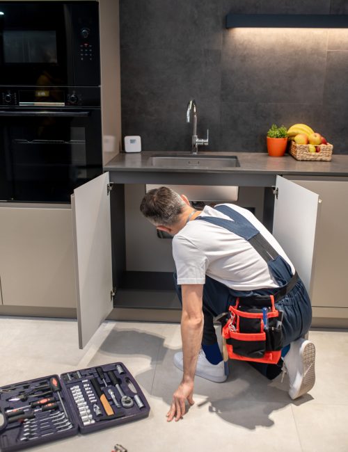 Searching for problem. Back view of man in white tshirt and blue overalls with tools on his belt crouching peeking under sink in beautiful modern kitchen